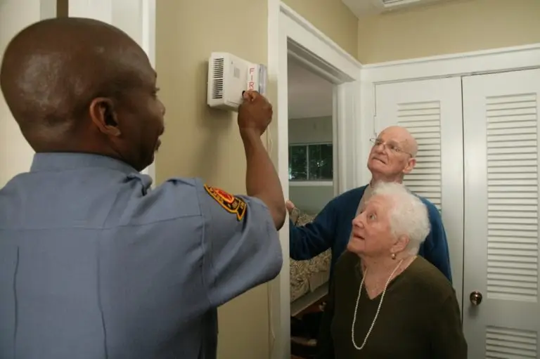 A fire safety officer installs a smoke detector in a home while an elderly couple looks on with interest.