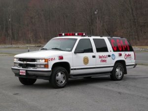 A white SUV with red and black markings, labeled "C 6-1," is parked in an open area. It has emergency lights on top and various logos on the sides. The background features a wooded area.