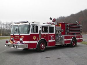 A large red and white fire truck is parked on gray pavement. It has various equipment on its sides and a ladder on top. The background shows a cloudy sky and leafless trees on a hill.