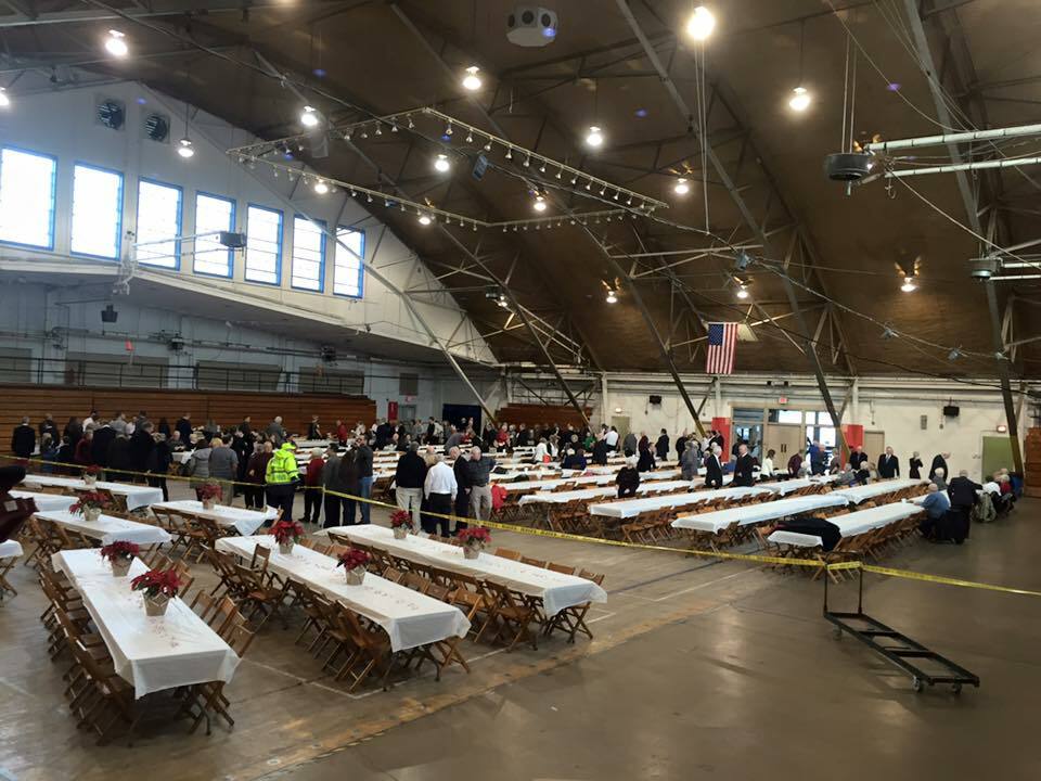 A large indoor hall with rows of long tables and chairs set up, covered with white tablecloths. People are gathered at one side of the room. The ceiling features exposed beams and lights. An American flag is hanging on the wall.