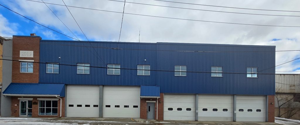 A building with a blue and red exterior, featuring four large white garage doors. Several windows are aligned at the top. A number "2919" is visible on the brick section. The sky is cloudy, with a light dusting of snow on the ground.