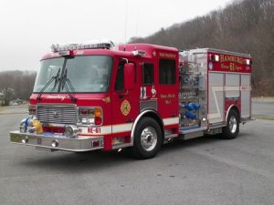 A red fire truck labeled "HAMBURG" with number 61, parked on a paved area. It has multiple compartments and equipment. The background features a cloudy sky and a tree-lined hill.