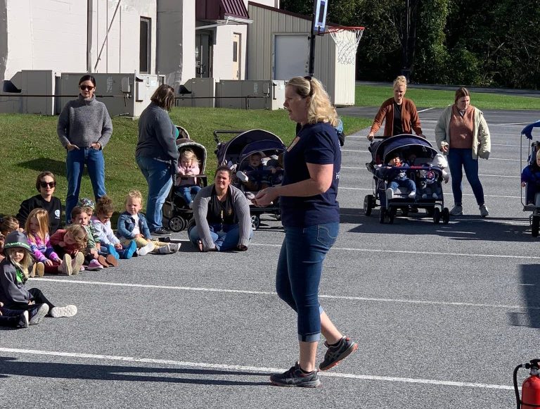 A woman is holding a microphone and speaking to a group of children sitting outdoors. Several strollers with infants are nearby. Adults are standing or sitting with the children on a sunny day. A fire extinguisher is on the ground.