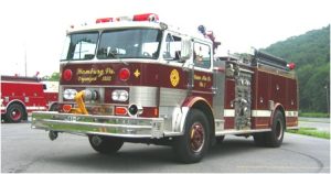 A vintage red and white fire truck is parked outdoors. It has multiple hoses and equipment on its side and "Hamburg, Pa. Organized 1832" written on the front. Trees and a hill are visible in the background.