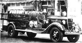 A vintage black and white photo of an old-fashioned fire engine with a ladder on one side. The truck has classic round headlights and a hose reel mounted on the back. Buildings and awnings are visible in the background.