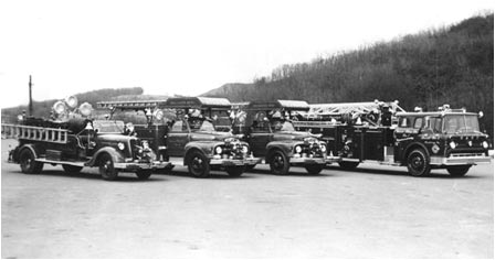 Black and white image of four vintage fire trucks parked in a row on a paved area. The trucks have ladders and equipment on top. Trees and hills are visible in the background.